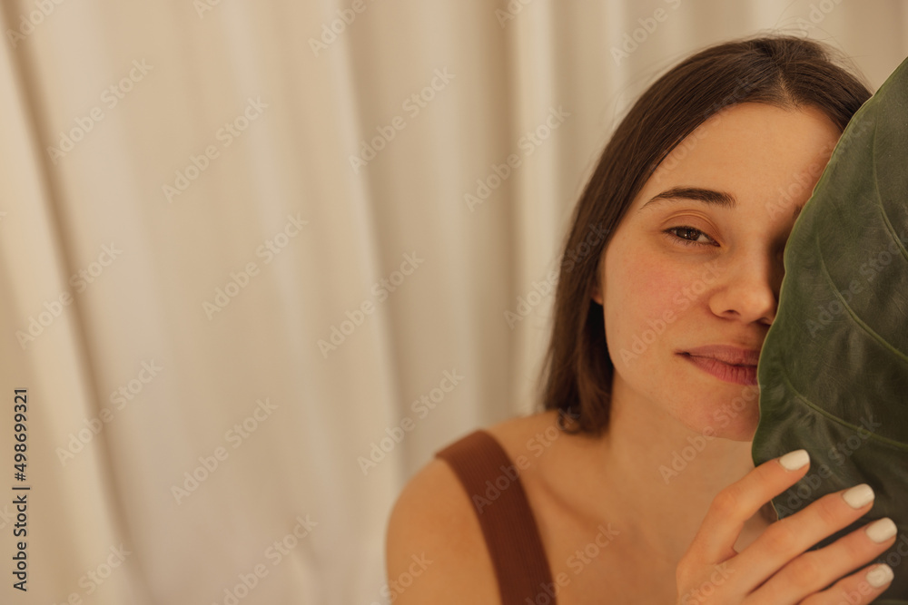 Close up beautiful caucasian young woman with natural makeup and clean fresh healthy skin covers her face with palm leaf. Brunette looks at camera, posing against background of white wall. Spa concept Close up beautiful caucasian young woman with natural makeup and clean fresh healthy skin covers her face with palm leaf. Brunette looks at camera, posing against background of white wall. Spa concept
