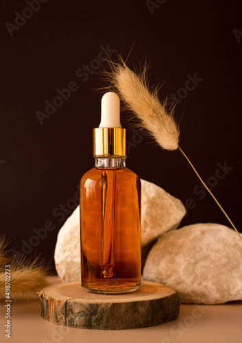 Transparent glass cosmetic dropper standing on wooden podium near stones and dry flowers on dark beige background. Beauty composition. Concept of natural and original skincare products presentation. 
