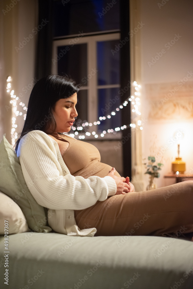 Side view of pretty pregnant woman sitting on bed. Barefoot woman in dress and cardigan touching big belly. Pregnancy, expectation concept Side view of pretty pregnant woman sitting on bed. Barefoot woman in dress and cardigan touching big belly. Pregnancy, expectation concept