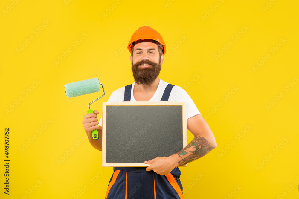 smiling man decorator in helmet and work clothes hold paint roller and blackboard smiling man decorator in helmet and work clothes hold paint roller and blackboard