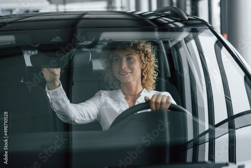 Sitting at driver's seat. Woman with curly blonde hair is in autosalon