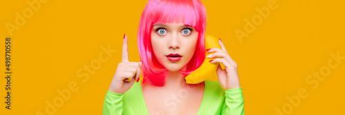 Surprised young sexy woman on pink wig call by banana and shows up finger on yellow background