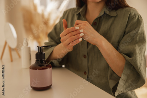 Close-up of young caucasian girl applying moisturizer on her hands while sitting at table. Lady with healthy skin uses skin care cosmetics. Women's beauty concept