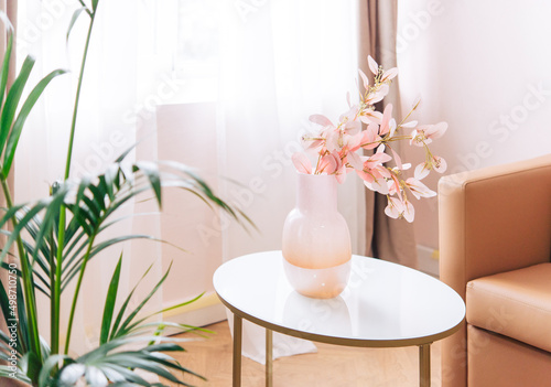Soft home decor, glass jug, vase with white and pink beautiful flowers against window and curtains and table top. interior. 