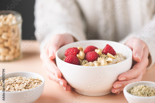 Oatmeal bowl with raspberry, cashew nuts and hemp seeds in female's hands. Clean eating, dieting concept