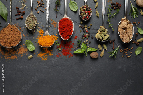 Different herbs and spices with spoons on black table, flat lay. Space for text