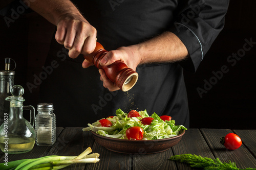 Professional chef adds peppers to a fresh vegetable salad in kitchen. Close-up of cook hands holding a mill. Cooking healthy and tasty food