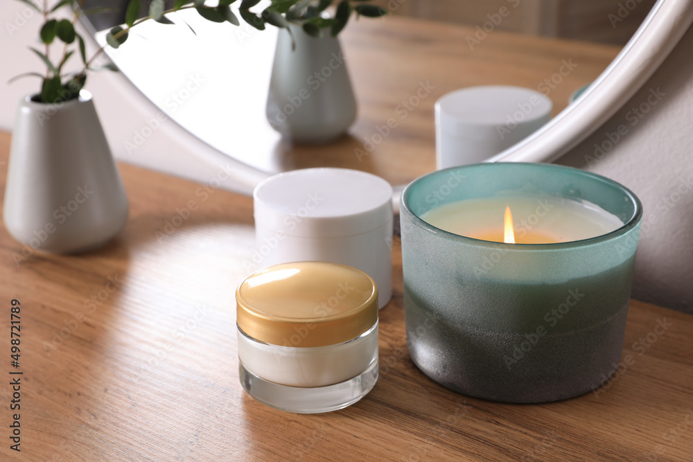 Burning candle, cosmetic products and vase on wooden dressing table, closeup