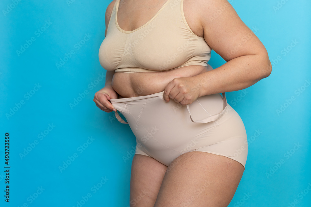 Plus size woman in beige underclothes pulling shapewear panties over belly closeup. Tighten figure and wear lifting underwear for weight loss, blue background. Concept of diet and fighting overweight. - obrazy, fototapety, plakaty 