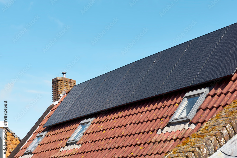 A row of dark solar panels on a house roof with orange tiles against a blue sky.     