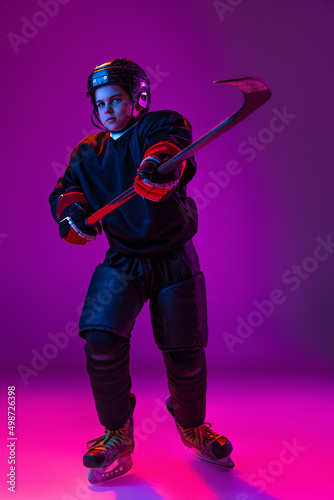 Portrait of little boy, child, hockey player in special uniform posing with stick isolated over purple background in neon light. Playful childhood
