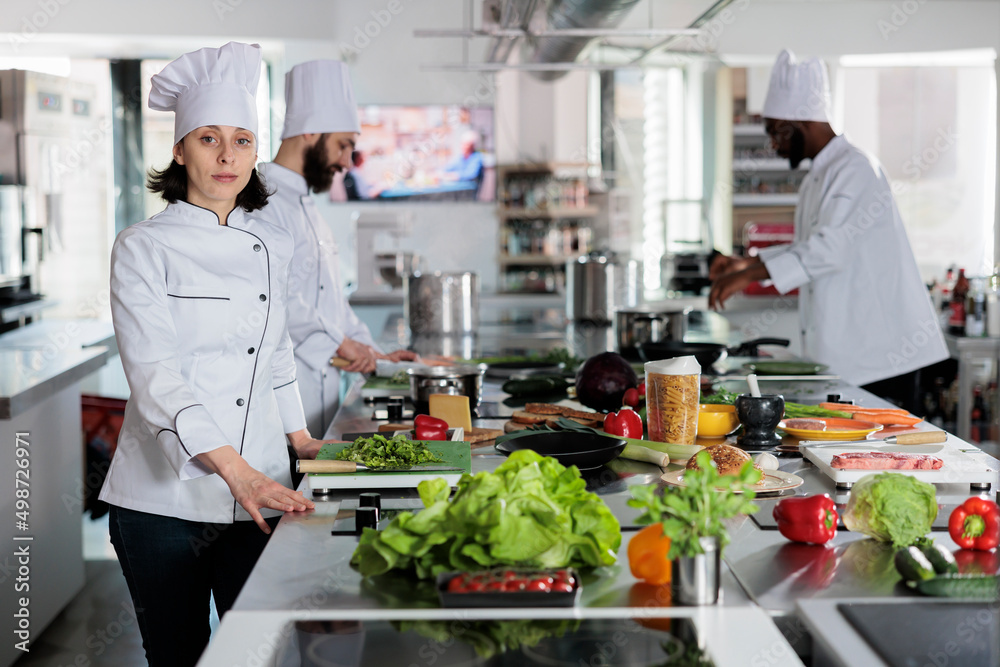 Head cook preparing spices and ingredients for dinner dish while looking confident at camera. Sous chef standing in restaurant professional kitchen space with arms crossed.