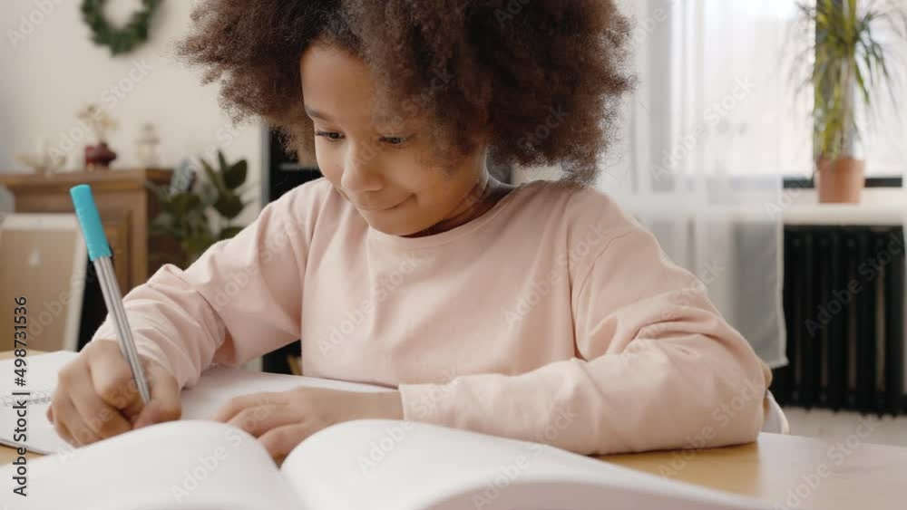 Zoom in portrait of cute concentrated little african american girl writing in copy book, studying at home, slow motion Zoom in portrait of cute concentrated little african american girl writing in copy book, studying at home, slow motion