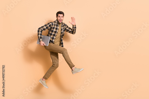 Full length body size view of attractive cheery guy jumping holding laptop waving hi isolated on beige pastel color background