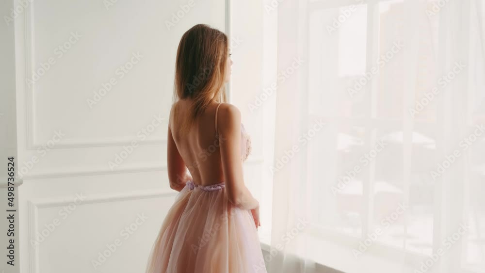 Portrait adult blonde girl princess standing by large panoramic window. Young woman enjoys view. Open sexy back, blonde hair, silk curtain flying fluttering in wind. Pink dress ball gown. White room Portrait adult blonde girl princess standing by large panoramic window. Young woman enjoys view. Open sexy back, blonde hair, silk curtain flying fluttering in wind. Pink dress ball gown. White room