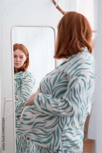 pregnancy redhead woman in pajamas looking at mirror at her belly 