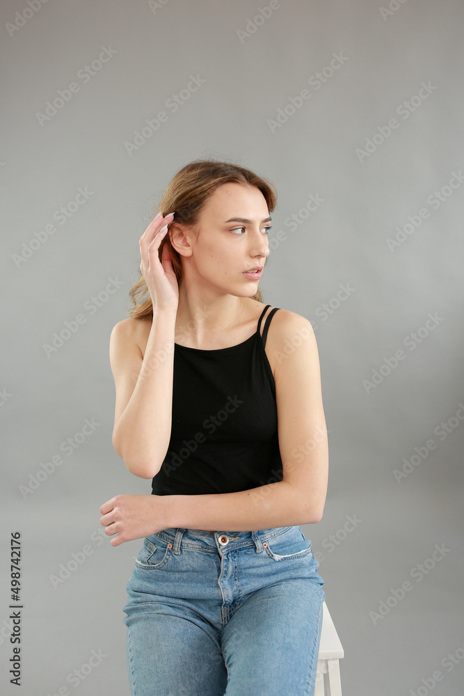 Beautiful girl model appearance in light jeans and black T-shirt on a gray studio background. Girl model sitting on a white chair. Beautiful blonde shows poses for a photo shoot
