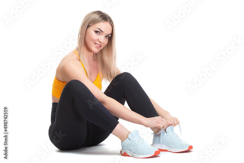 A beautiful, athletic, slim, smiling and cheerful woman in an orange top and black sweatpants is sitting and tying her shoelaces on sports sneakers. Lifestyle concept with sports and gym, healthy