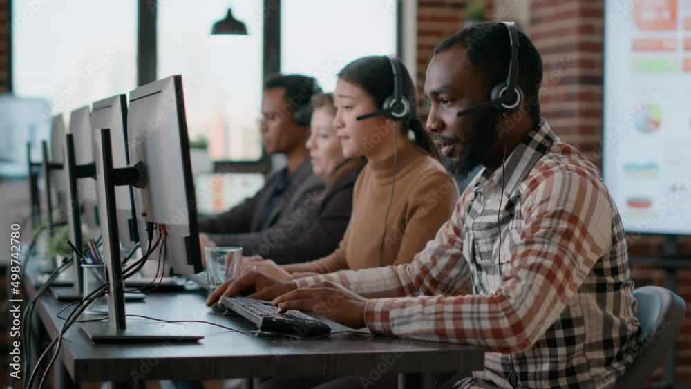 African american man working at call center office to help people with telemarketing assistance. Male employee using headphones and microphone at customer care service. Tripod shot. African american man working at call center office to help people with telemarketing assistance. Male employee using headphones and microphone at customer care service. Tripod shot.