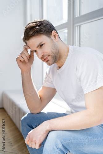 positive and dreamy man holding hand near head while sitting on windowsill.