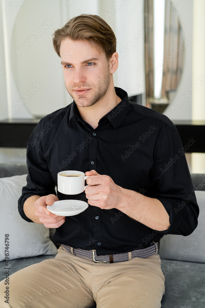 man in black shirt sitting with coffee cup and saucer on couch in living room. man in black shirt sitting with coffee cup and saucer on couch in living room.