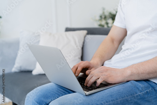 partial view of man sitting on sofa at home and typing on laptop.