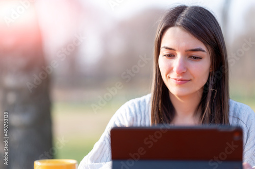Portrait of a smiling young beautiful woman working remotely in a park. In front of her, her tablet and her yellow cup of coffee. Beautiful spring day. Remote work outdoors concept.