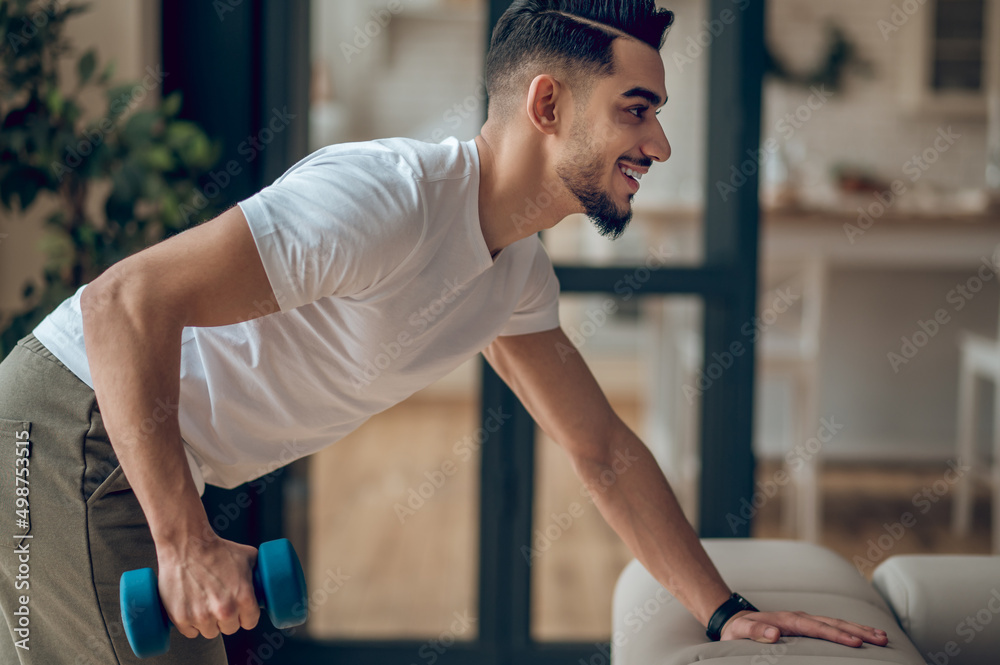 Fototapeta premium Young dark-haired man exercising with dumbbells and looking contented
