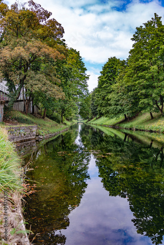 Calm river water reflects the surrounding green grass and coloured autumn trees