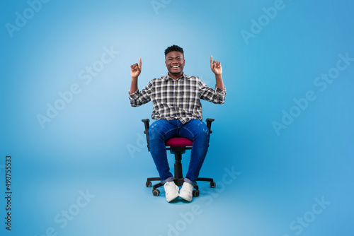 Handsome young black man pointing two fingers up at empty space, sitting on chair on blue studio background, mockup