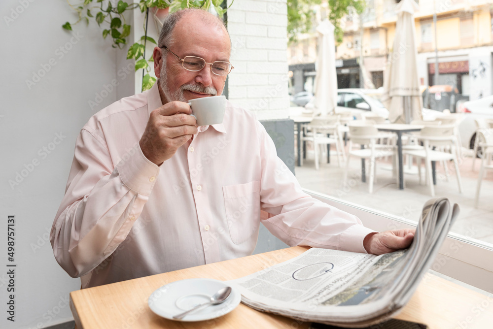a smiling older retired man with glasses reading a daily newspaper sitting in a coffee shop while drinking his coffee a smiling older retired man with glasses reading a daily newspaper sitting in a coffee shop while drinking his coffee