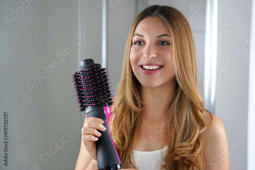Beatiful girl holds round brush hair dryer to style hair at home. Young woman showing salon one-step hair dryer and volumizer.