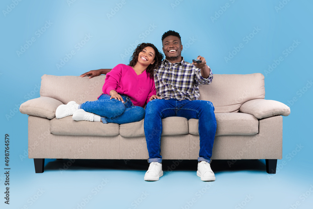 Excited young black couple with remote control sitting on couch and watching TV against blue studio background Excited young black couple with remote control sitting on couch and watching TV against blue studio background