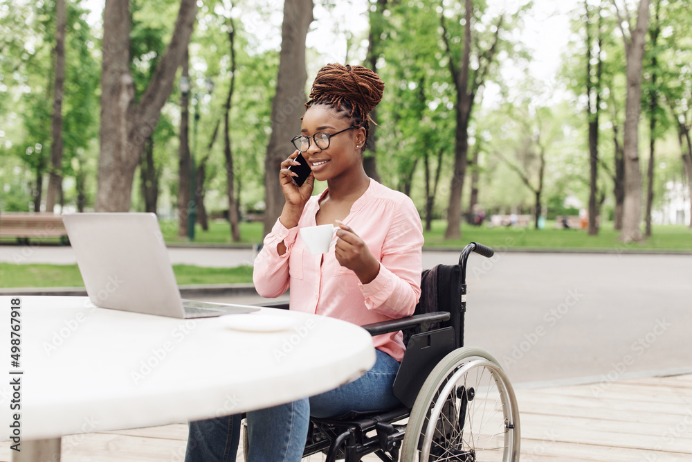 Young black woman in wheelchair working online, using laptop, speaking on smartphone and enjoying coffee at outdoor cafe Young black woman in wheelchair working online, using laptop, speaking on smartphone and enjoying coffee at outdoor cafe
