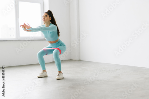 Young athletic woman doing squats using rubber resistance band at gym
