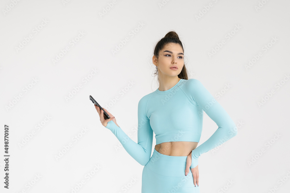 Young white fitness woman holding mobile phone in sportswear with ponytail standing over white wall background Young white fitness woman holding mobile phone in sportswear with ponytail standing over white wall background