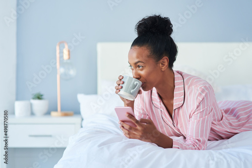 Nothing tastes better than morning gossip. Shot of an attractive young woman drinking coffee while using her cellphone in bed at home.