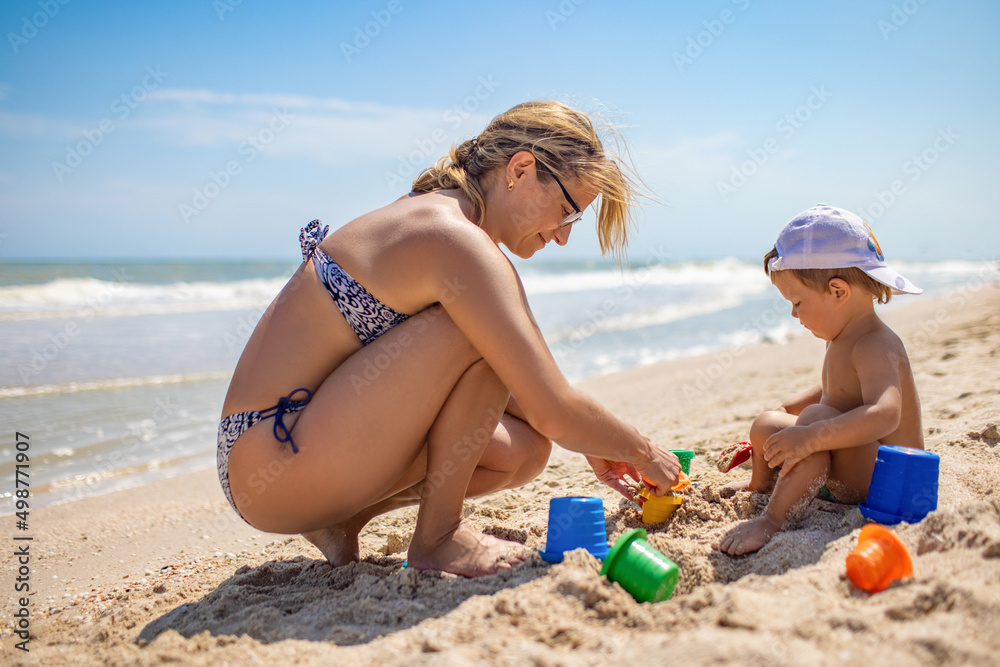 Mom with glasses plays with a little toddler sitting on the beach near the sea Mom with glasses plays with a little toddler sitting on the beach near the sea