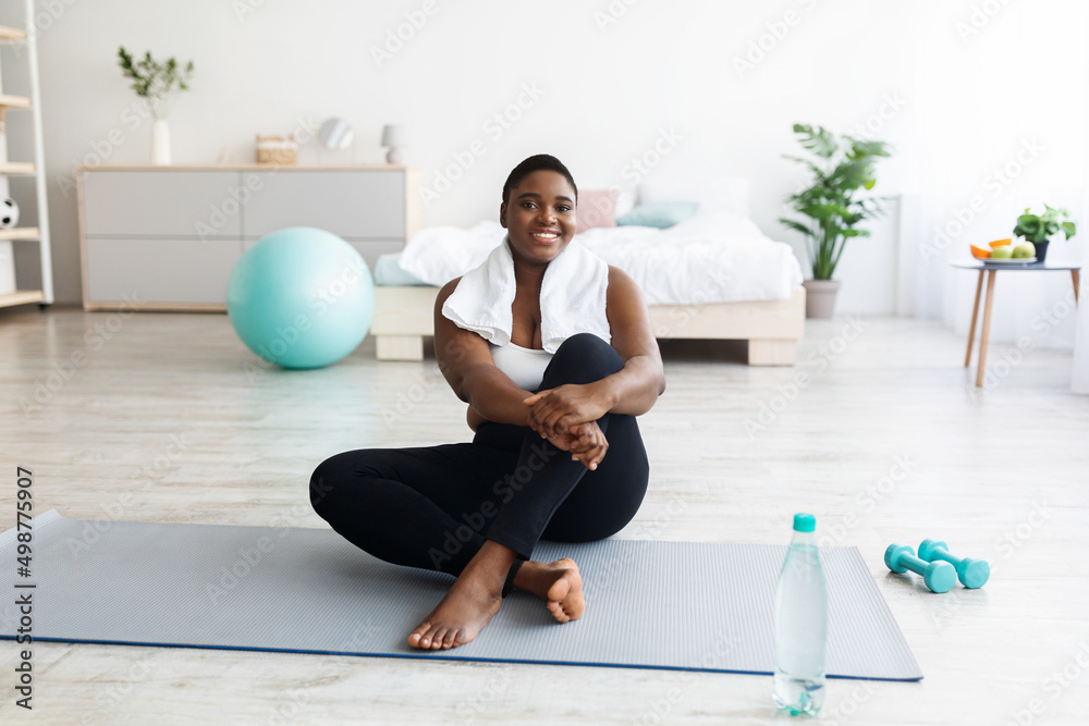Fototapeta premium Smiling overweight young black woman sitting on yoga mat, resting after home fitness, copy space