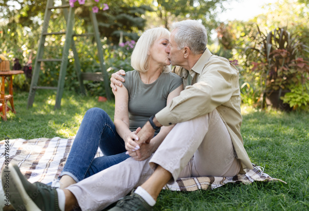 Married senior couple resting outdoors and kissing, enjoying holiday while sitting on blanket on grass in garden
