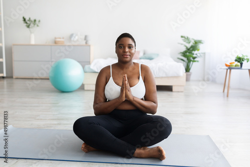 Overweight young black woman sitting on mat in lotus pose, making namaste gesture, meditating, practicing yoga at home