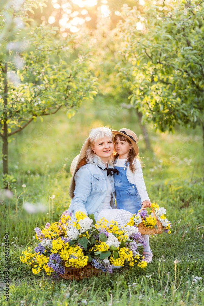 beautiful blonde woman and her little daughter are sitting in a spring garden with a large basket of flowers. beautiful blonde woman and her little daughter are sitting in a spring garden with a large basket of flowers.