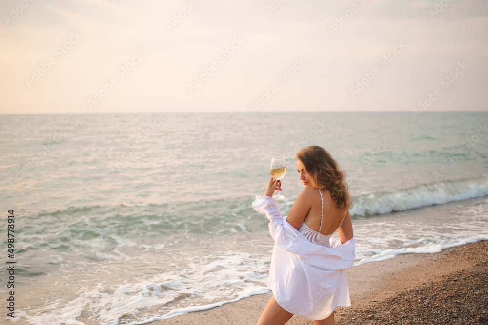 Beautiful blonde adult girl wear elegant swimsuit and white shirt resting over blue sea shore hold glass of wine over nature background outdoors. Summer vacation season. 20s. Stylish woman relax