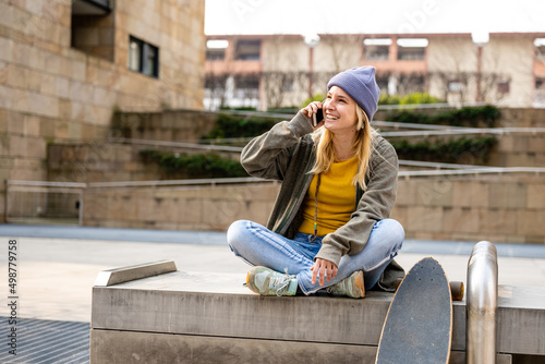 Young blonde woman with veri peri coolor hat talking with friend using smart phone, teenager female skater sitting on a bench in the city, copy space photo