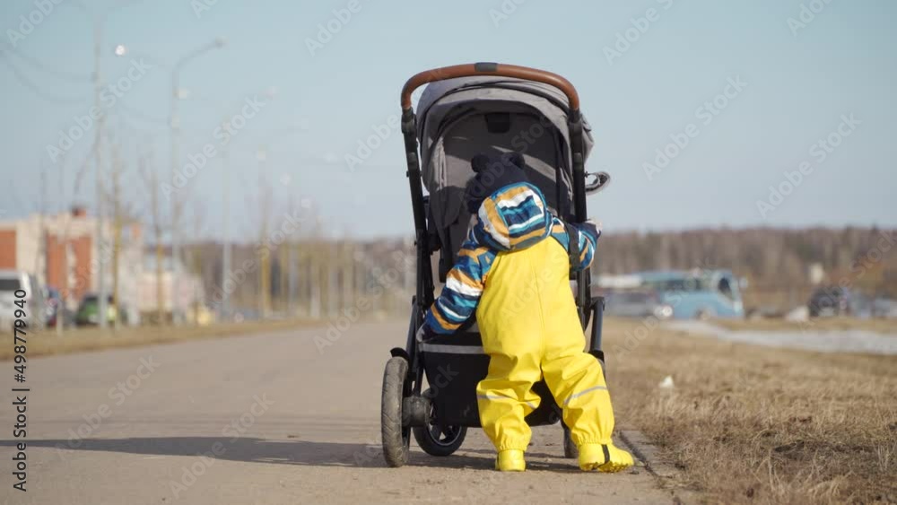 Mom with small cute child in yellow waterproof rain pants bibs for kids walking on the footpath, one year old baby boy learning to walk in the cold season. High quality 4k footage Mom with small cute child in yellow waterproof rain pants bibs for kids walking on the footpath, one year old baby boy learning to walk in the cold season. High quality 4k footage