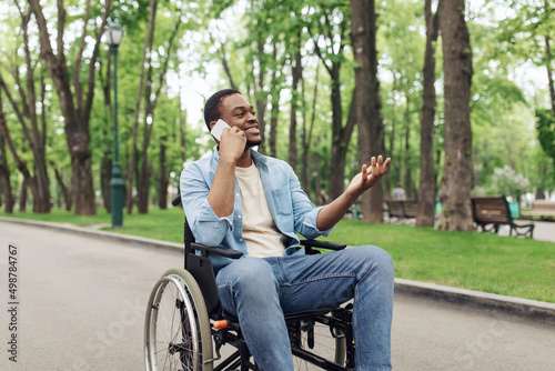 Young black guy in wheelchair having phone conversation at city park in summer