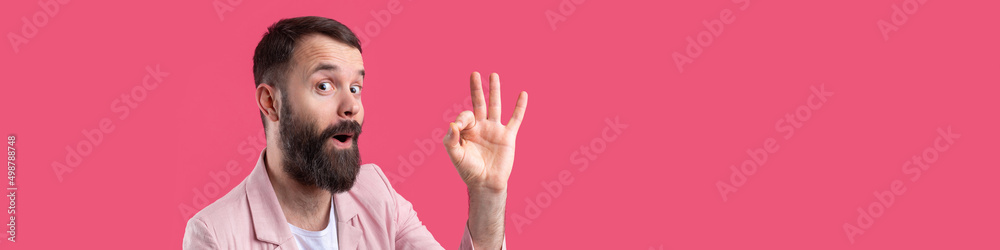 Fototapeta premium Portrait of a smiling young man in a pink jacket. Man showing ok gesture while standing in studio over isolated red background.