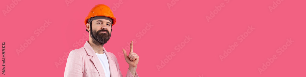 Portrait of young construction engineer wear orange hard hat, in a pink jacket standing on red studio background. The man points with his hand. Portrait of young construction engineer wear orange hard hat, in a pink jacket standing on red studio background. The man points with his hand.