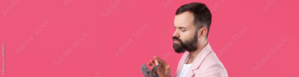 Fototapeta premium Portrait of a contented young businessman with a beard dressed in a pink jacket showing us dollar banknotes against a red studio background. Taste, smell of money.