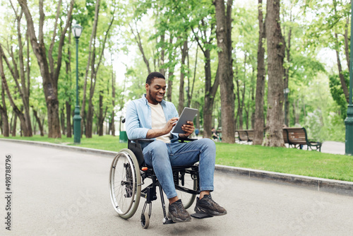 Positive young black guy in wheelchair using tablet computer, browsing web or watching video at urban park, copy space